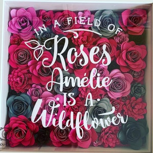 May include: A white frame filled with pink and gray paper flowers. The text "In a field of roses Amelie is a wildflower" is written in white on the flowers.