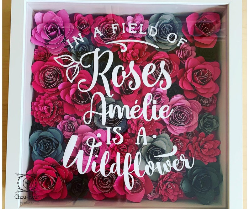 May include: A white frame filled with pink and gray paper flowers. The text "In a field of roses Amelie is a wildflower" is written in white on the flowers.