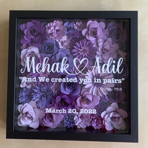 May include: A framed shadow box with a purple and white floral arrangement. The names "Mehak" and "Adil" are written in white with a heart symbol between them. The text "And We created you in pairs" is written in white below the names with the text "Quran - 78:8" below that. The date "March 20, 2022" is written in white at the bottom of the box.