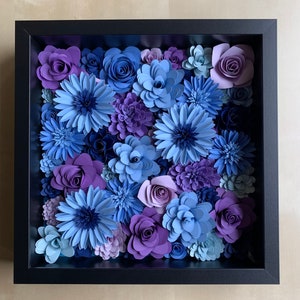 May include: A framed wall art piece featuring a variety of paper flowers in shades of blue, purple, and white. The flowers are arranged in a circular pattern and are surrounded by a black frame.
