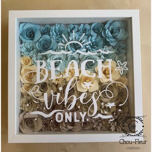 May include: A white shadow box filled with paper flowers in shades of blue, yellow, and brown. The flowers are arranged in a gradient pattern, with the blue flowers at the top and the brown flowers at the bottom. The words "Beach Vibes Only" are written in white on the front of the box.