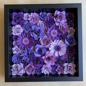 May include: A framed artwork featuring a variety of purple paper flowers in different sizes and shapes. The flowers are arranged in a dense cluster, creating a textured and dimensional effect. The frame is black and has a simple design.