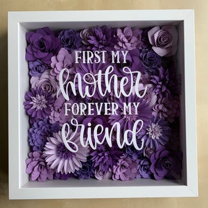 May include: A white frame with a purple paper flower arrangement. The flowers are in various shades of purple and the text "First my mother forever my friend" is in the center of the arrangement.