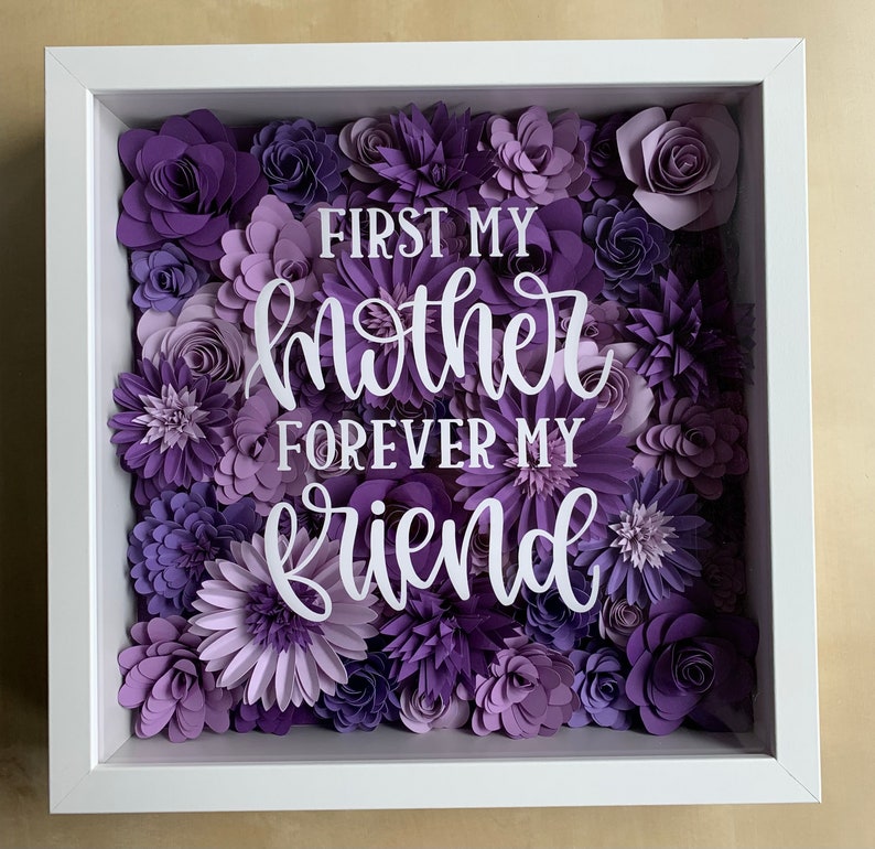 May include: A white frame with a purple paper flower arrangement. The flowers are in various shades of purple and the text "First my mother forever my friend" is in the center of the arrangement.