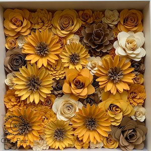 May include: A framed arrangement of paper flowers in shades of yellow, brown, and cream. The flowers are arranged in a circular pattern and are all different sizes and shapes. The frame is white and has a simple design.