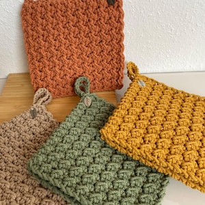 Könnte beinhalten: Eine Auswahl an handgefertigten Topflappen in Rost-, Beige-, Grün- und Goldtönen. Jeder quadratische Topflappen hat ein strukturiertes Stichmuster und eine Schlaufe zum Aufhängen. Ein Holzknopf ist an der Schlaufe befestigt.