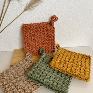Könnte beinhalten: Vier gehäkelte Topflappen in verschiedenen Farben: Braun, Grün, Gelb und Orange. Jeder Topflappen hat eine Schlaufe zum Aufhängen.