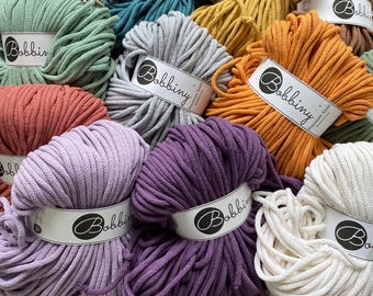 May include: A collection of multiple skeins of Bobbiny 5mm braided cord in various colors including sage green, light gray, mustard yellow, terracotta, lavender, deep purple, and off-white, each featuring a white paper label with the brand name.