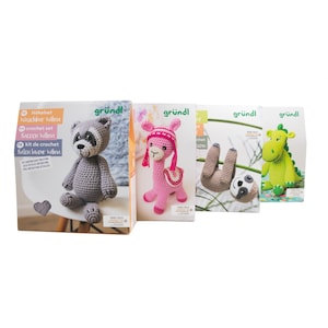 Könnte beinhalten: Eine Auswahl an Häkelsets mit verschiedenen Tierdesigns. Die Sets beinhalten einen Waschbären, ein rosa Lama, ein Faultier und einen grünen Dinosaurier. Jedes Set ist in einer Box mit dem "gründl"-Logo und Anleitungen verpackt.