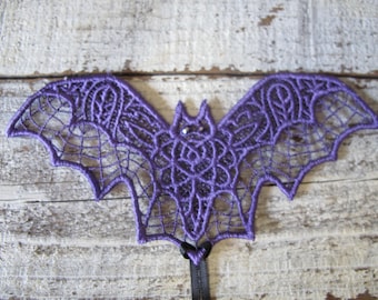 Puede incluir: Decoración de murciélago de encaje morado con un ojo de cuentas. El murciélago está intrincadamente diseñado con un patrón de telaraña y un cuerpo en forma de rosa.