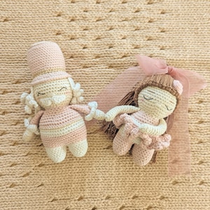 May include: Two handmade crochet dolls, one shaped like a nutcracker in a pink top hat and the other like a ballerina with brown hair and a pink ribbon, both featuring embroidered closed eyes and soft pastel yarn construction.