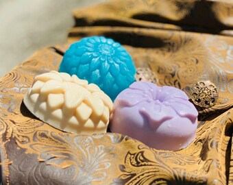 May include: Three handmade soap bars with floral designs. The soaps are in shades of blue, white, and purple. The soaps are sitting on a brown patterned fabric.