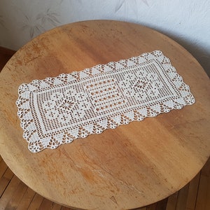 May include: White crocheted table runner with a delicate floral pattern. The runner is laid on a wooden table.