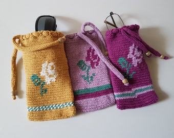 May include: Three crocheted eyeglass cases with floral designs. The cases are in shades of yellow, pink, and purple. Each case has a drawstring closure and a beaded tassel.