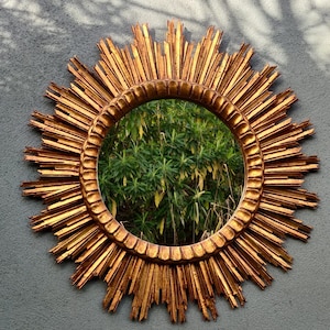 May include: A large gold sunburst mirror with a round mirror in the centre. The mirror reflects a green bush.
