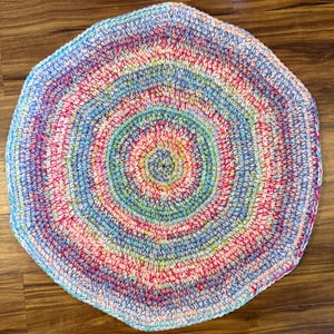 May include: A handmade, colorful crochet rug with a spiral design. The rug features a mix of pastel colors, including pink, blue, green, and yellow. The rug is round with a hexagonal shape and is made of soft yarn.