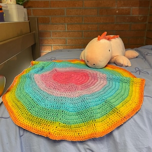 May include: A rainbow crochet baby blanket with a pink, blue, green, yellow, and orange color gradient. The blanket is on a light blue bedspread with a plush toy in the background.