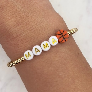 May include: A gold beaded bracelet with white letter beads spelling "MAMA" and a small orange basketball charm.