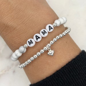 May include: Two bracelets on a wrist. One bracelet is made of white beads with the word "MAMA" spelled out in black letters. The other bracelet is made of silver beads with a small silver heart charm.