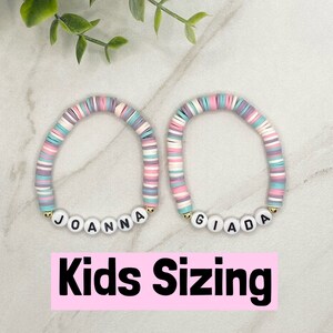 May include: Two colorful beaded bracelets with the names "JOANNA" and "GIADA" spelled out in white letters. The bracelets are on a white marble surface. Kids Sizing.