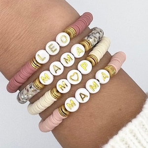 May include: A set of four bracelets with white beads and gold lettering. The bracelets spell out "LEO", "MAMA", "MAMA", and "MAMA". One of the bracelets has a heart charm.