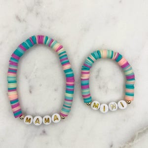 May include: Two colourful beaded bracelets with the words "MAMA" and "MINI" spelled out in white beads. The bracelets are made of pastel pink, blue, and green beads.