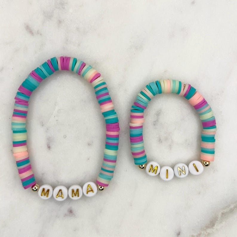 May include: Two colourful beaded bracelets with the words "MAMA" and "MINI" spelled out in white beads. The bracelets are made of pastel pink, blue, and green beads.