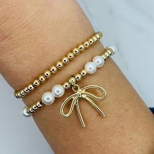 May include: Two gold bracelets with white pearls and a gold bow charm. The bracelets are stacked on a person's wrist.