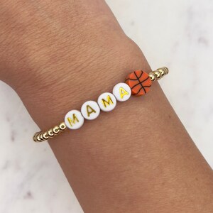 May include: A gold beaded bracelet with white letter beads spelling "MAMA" and an orange basketball charm.
