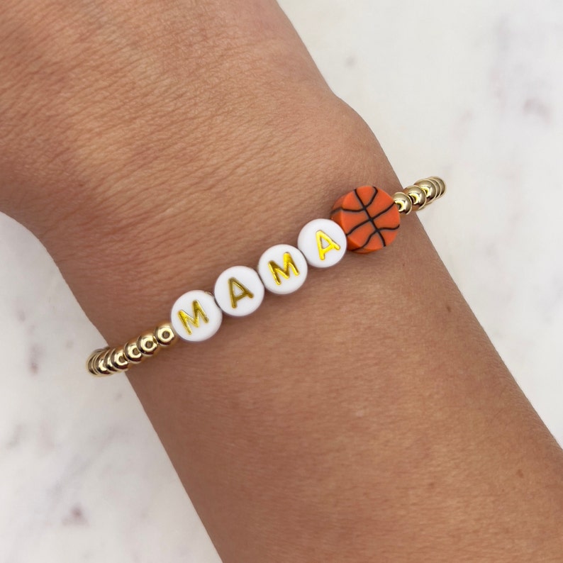 May include: A gold beaded bracelet with white letter beads spelling "MAMA" and an orange basketball charm.