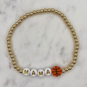 May include: Gold beaded bracelet with white beads spelling "MAMA" and an orange basketball charm.