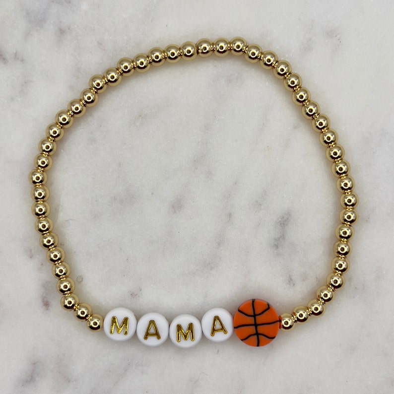 May include: Gold beaded bracelet with white beads spelling "MAMA" and an orange basketball charm.