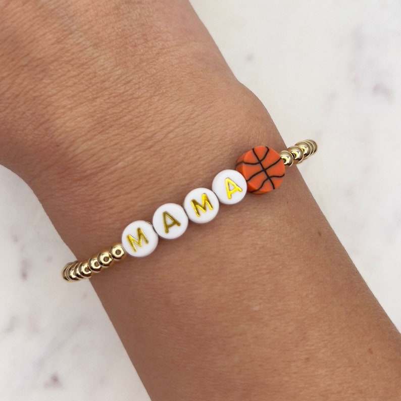 May include: A gold beaded bracelet with white letter beads spelling "MAMA" and a small orange basketball charm.