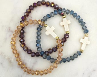 May include: Three beaded bracelets in shades of gold, purple, and blue. Each bracelet features faceted beads and a white cross charm. The bracelets are arranged in a circular pattern on a marble surface.