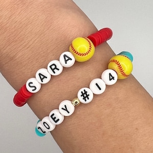 May include: Two beaded bracelets. One red bracelet spells "SARA" with a yellow softball bead. The other bracelet is turquoise and spells "ZOEY #14" with a yellow softball bead. The bracelets are on a wrist.
