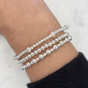 May include: Three silver beaded bracelets stacked on a wrist. The bracelets are made of small, round beads with a slightly textured surface.