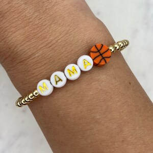 May include: A gold beaded bracelet with white letter beads spelling "MAMA" and an orange basketball charm.