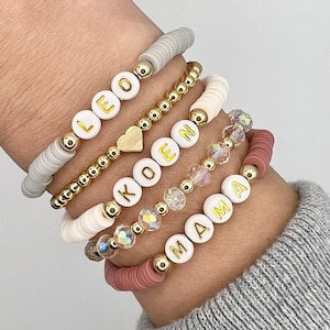May include: A set of six bracelets featuring white beads with gold lettering spelling out the names "LEO", "KOEN", "MAMMA", and a heart charm. The bracelets are layered on a person's wrist, with a crystal beaded bracelet and a pink beaded bracelet.