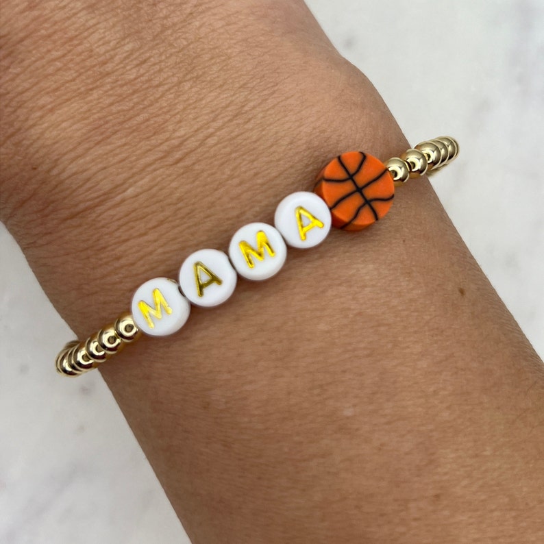 May include: A gold bracelet with white beads that spell out "MAMA" and a small orange basketball charm.