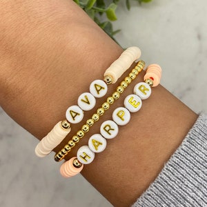 May include: Three personalized bracelets stacked on a wrist. The top bracelet is cream-colored with the name "AVA" spelled out in gold letters. The middle bracelet is gold beaded. The bottom bracelet is peach-colored with the name "HARPER" spelled out in gold letters.