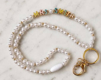 May include: A beaded lanyard featuring white faux pearls and small gold-tone spacer beads. The strand includes colorful letter beads spelling out MRS SMITH. A gold-tone metal keyring and lobster clasp are attached to one end, with a white plastic breakaway clasp.