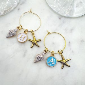 May include: Two gold-colored wine charms with seashell, starfish, and initial pendants. One charm has a white initial "H" pendant, and the other has a blue initial "A" pendant. The charms are designed to hang on the rim of a wine glass.
