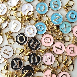 May include: A collection of gold-colored lobster clasp charms with enamel letter pendants in various colors, including white, black, pink, and blue. Each charm features a different letter of the alphabet.