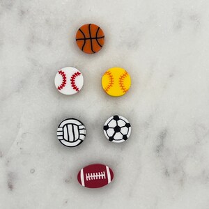 May include: Six different sports balls, including a basketball, baseball, softball, volleyball, soccer ball, and football. The balls are all white with black and red markings.