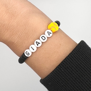 May include: A black and white beaded bracelet with the name "GIADA" spelled out in white beads. A yellow softball charm is attached to the bracelet.