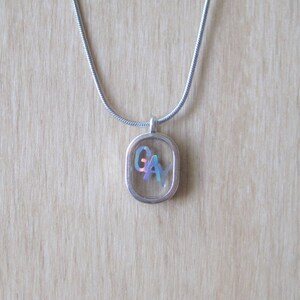 May include: A silver chain necklace with a square pendant featuring the letters "GAY" in iridescent colors.