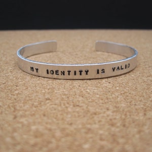 May include: A silver-colored cuff bracelet with the stamped text "MY IDENTITY IS VALID". The open-ended bracelet is displayed on a light brown surface, with a dark background.