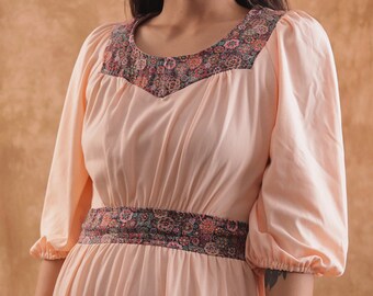May include: A light peach-colored dress with a floral patterned neckline and waist. The dress features puffed sleeves and a flowing skirt. The floral pattern includes shades of pink, green, and brown. The dress is made of a soft, flowing fabric.