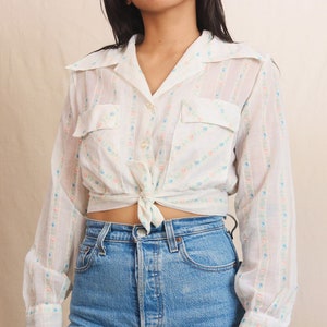 May include: White button-up shirt with a floral pattern and two chest pockets, tied at the waist. The shirt is paired with light blue denim jeans. The shirt features a delicate pattern of pink and blue flowers.