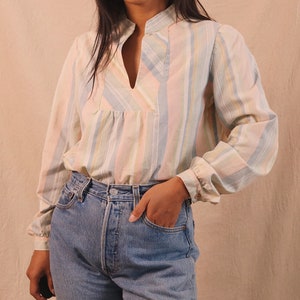 Peut inclure: Un chemisier &agrave; rayures pastel avec des manches longues et un d&eacute;collet&eacute; en V. Le chemisier pr&eacute;sente des rayures verticales dans les tons de rose, bleu, jaune et blanc. Le mod&egrave;le porte un jean en denim bleu.